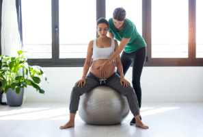 Embarazada sentada sobre pelota de Pilates con su entrenadora
